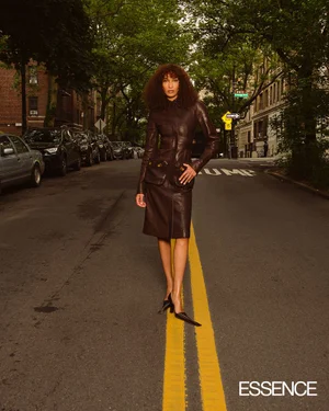 Model standing on a city street between parked cars wearing a brown leather coat and dark shoes