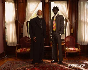 Two men standing in a wood-paneled room with ornate windows, wearing structured black and grey suits for Essence Magazine.