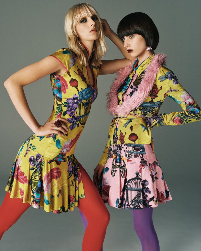 Two fashion models in colorful, patterned dresses, orange and purple tights pose in a studio with a neutral gray background.