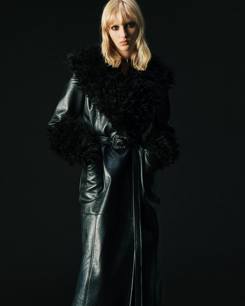 Blonde woman in a black, faux-fur-trimmed, belted, full-length leather coat poses against a dark background in a fashion editorial shot.
