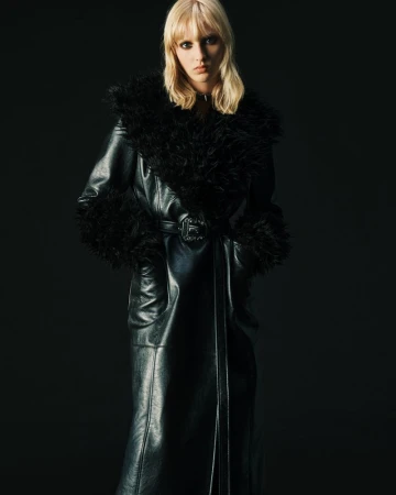 Blonde model in a belted black leather coat with voluminous faux fur collar and cuffs against a black studio background.