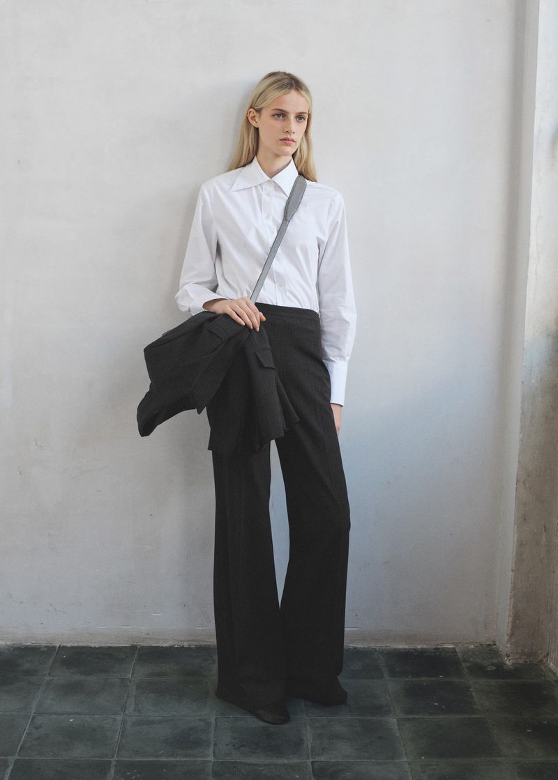 Fashion shot of a blond-haired woman in a white shirt and dark pants, carrying a black jacket on her shoulder against a neutral wall.