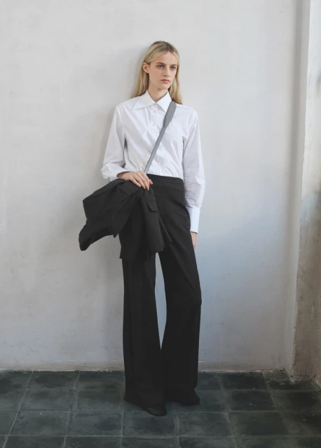 Fashion shot of a blond-haired woman in a white shirt and dark pants, carrying a black jacket on her shoulder against a neutral wall.