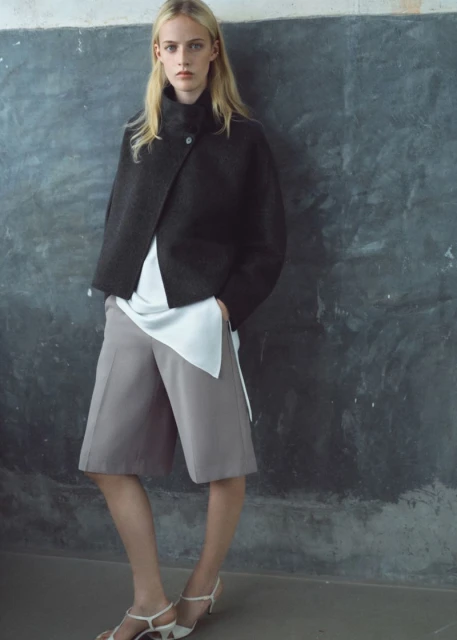 Blonde woman poses in a dark grey jacket, white top, and grey shorts against a textured grey wall in a studio-lit fashion shot.