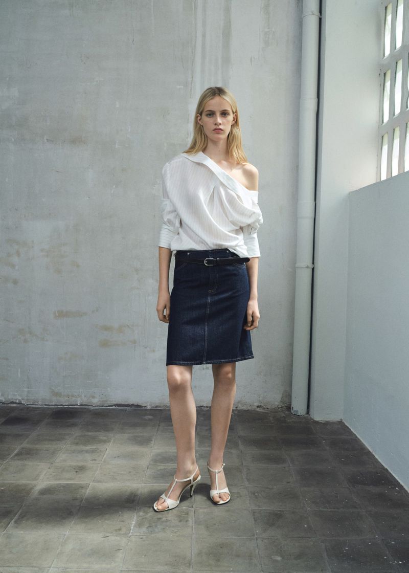 A woman poses in a white, off-the-shoulder shirt and a dark denim skirt with heeled sandals in a minimalist studio setting with natural light.