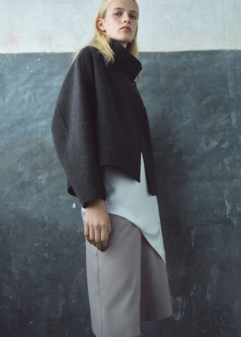 Fashion model poses in a grey jacket and culottes against a textured wall in a studio. Elegant minimalist design.