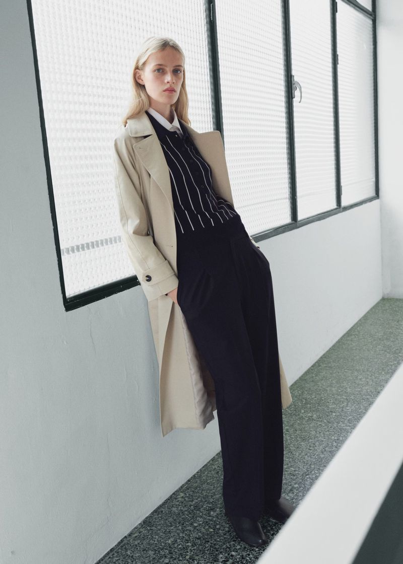 Elegant woman in trench coat poses indoors by window wearing pinstripe sweater, black wide-leg pants, and black heeled shoes in a studio setting.
