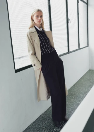 Elegant woman in trench coat poses indoors by window wearing pinstripe sweater, black wide-leg pants, and black heeled shoes in a studio setting.