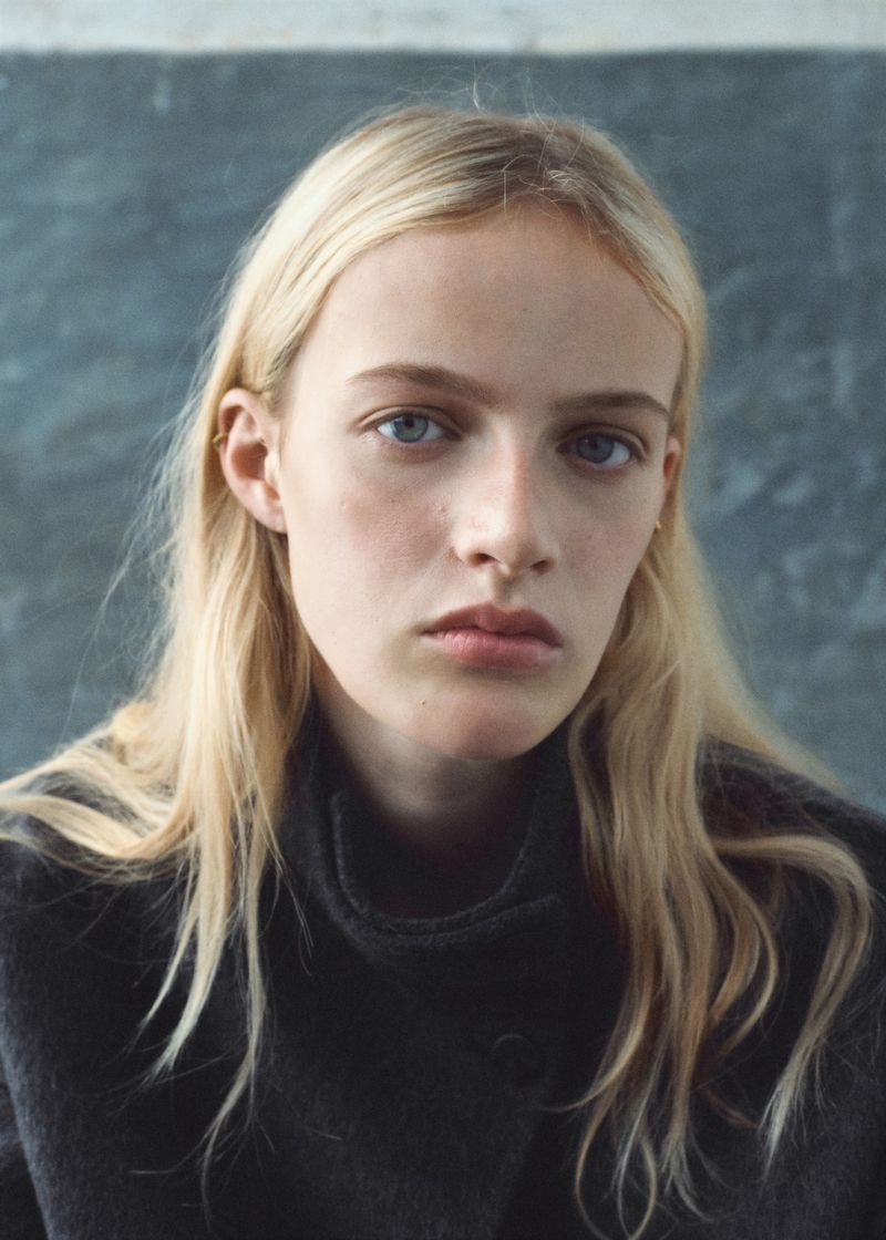 Close-up portrait of a blonde woman with blue eyes wearing a charcoal grey top, against a neutral grey background in soft light.