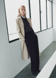 Model leaning against a wall wearing a beige trench coat, dark striped polo, and black trousers