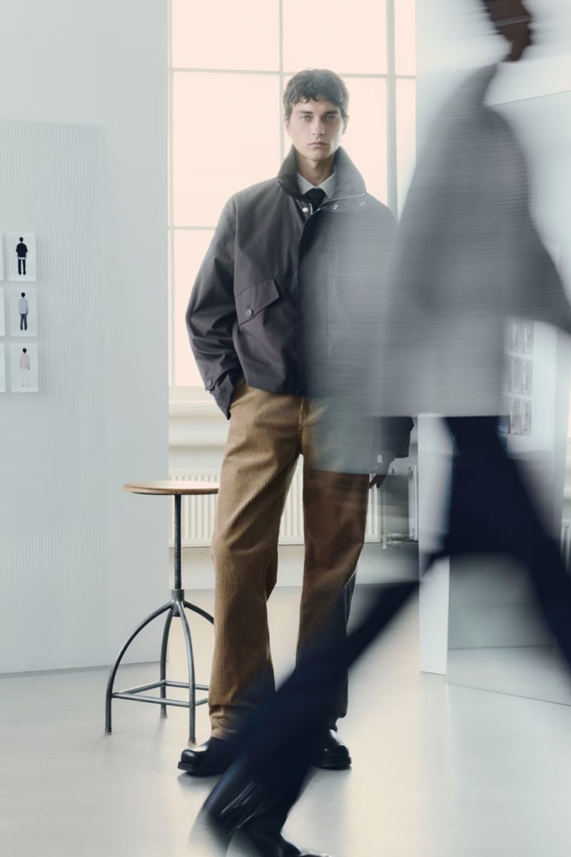 Young man posing in a gray jacket and brown trousers indoors, standing next to a small round table, with diffused light and motion blur.