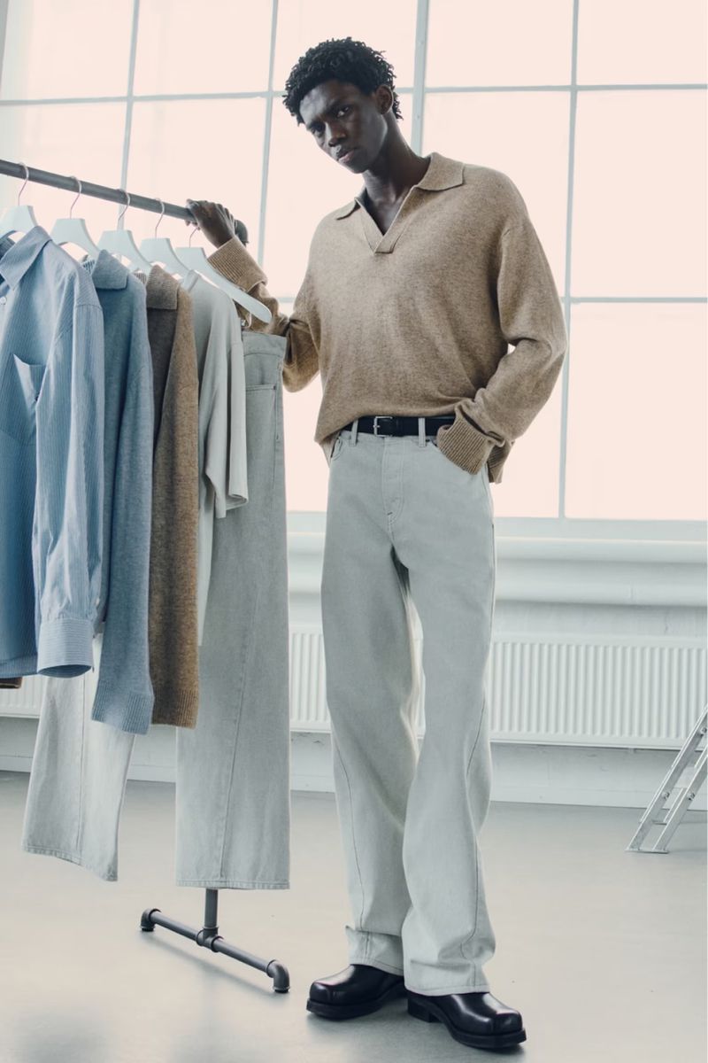 An African-American man poses indoors wearing a tan collared shirt and white pants, with a rack of clothes behind him.