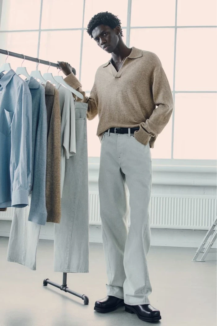 Model in camel knit polo shirt and light grey trousers leaning on a clothing rack in a bright studio.