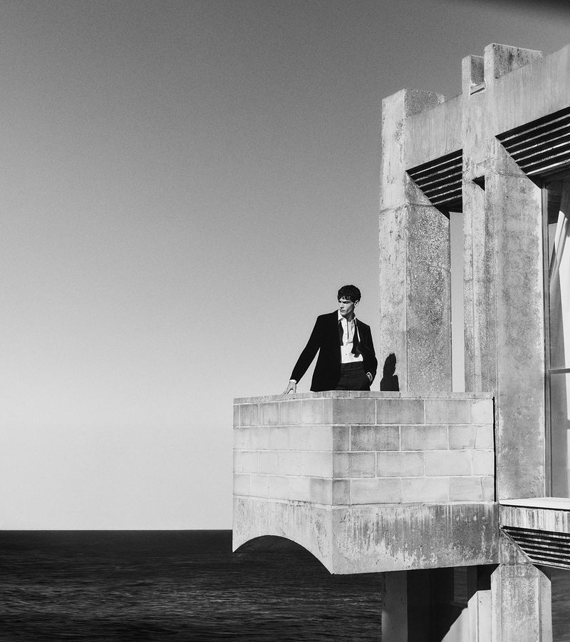 Monochrome portrait of a male model in a suit standing on a concrete balcony overlooking the ocean against a clear sky.