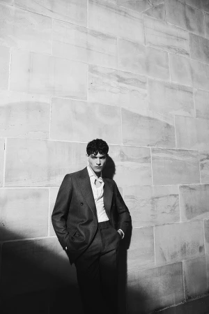 Monochrome portrait of a male model in a suit leaning against a textured stone wall, eyes forward, in fashion-editorial style.