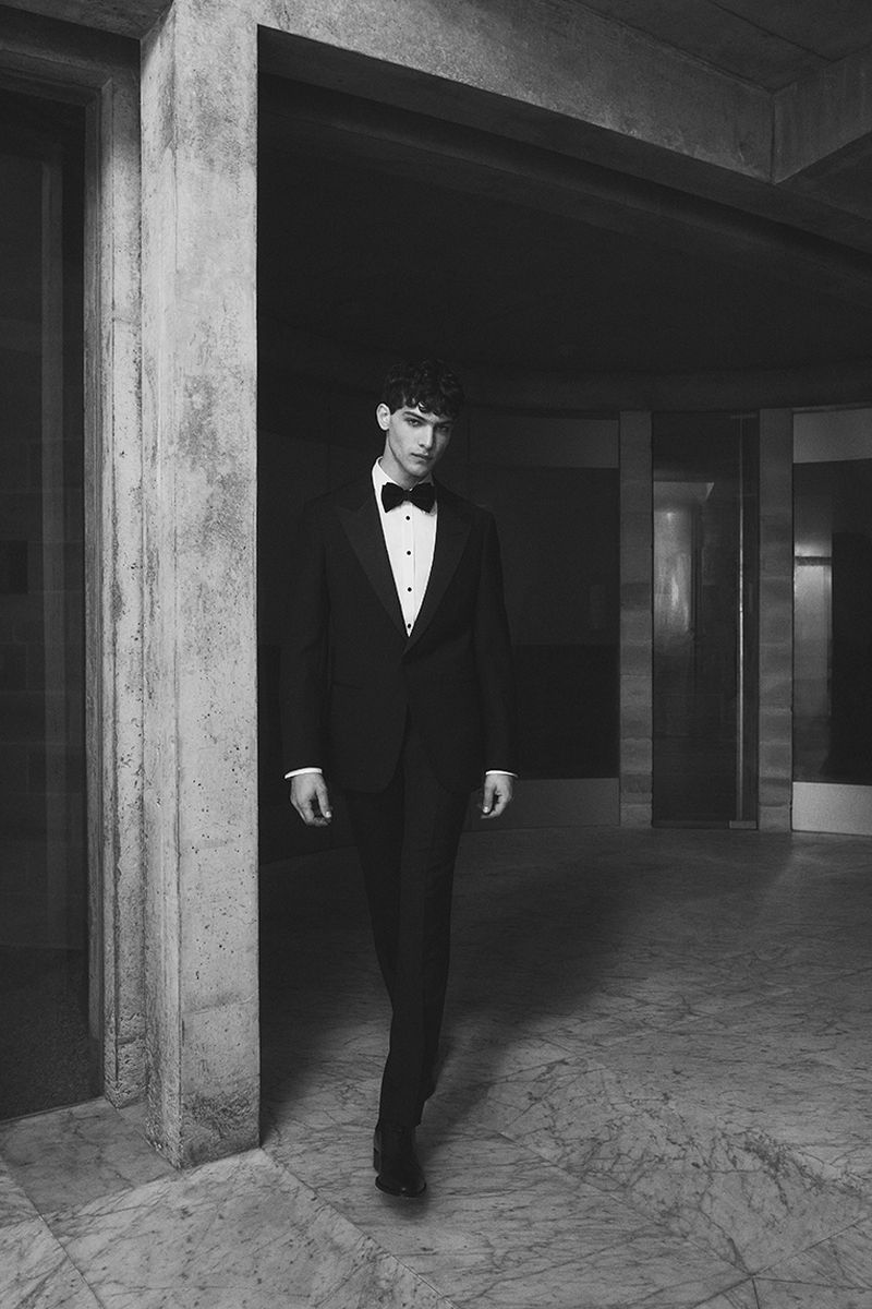 Monochrome portrait of a man in a black tuxedo with a bow tie, walking in an indoor space with marble flooring and concrete walls.