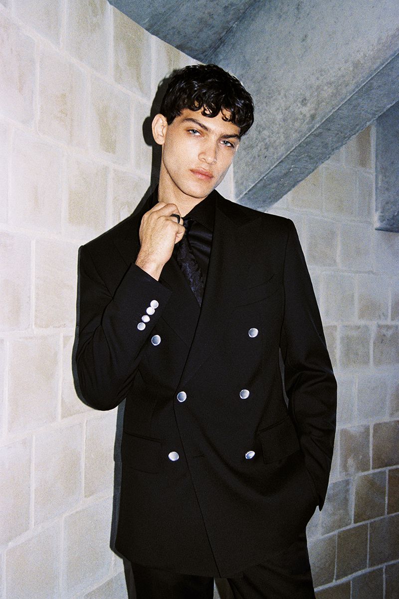 Man in a black double-breasted suit standing indoors against a textured wall, posing in formal attire with a dark shirt and tie.