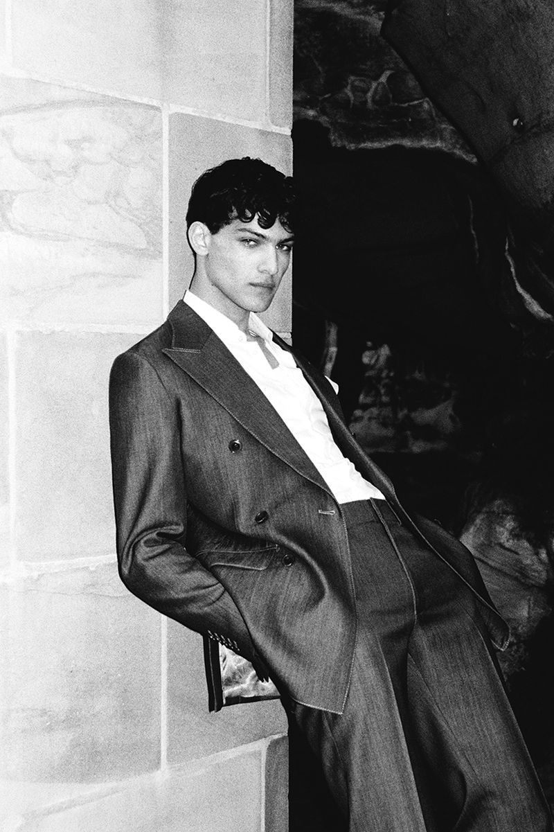 Black and white portrait of a young man in a suit leaning against a stone wall with a dark cavernous space in the background.