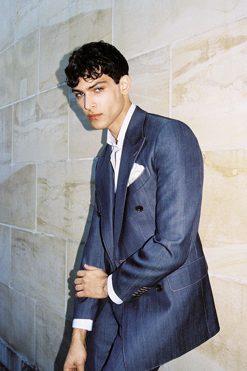 Young man poses in blue double-breasted suit with white pocket square against a light stone wall in a fashion photography shot.