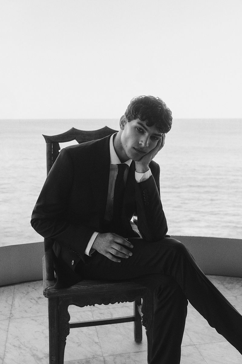 Young man in a black suit sits thoughtfully on a chair against an ocean backdrop, showcasing formal wear and classic portraiture.