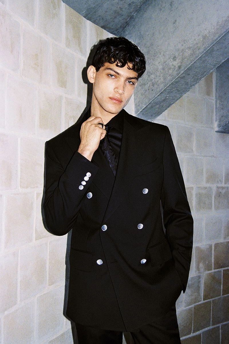 A man in a black double-breasted suit standing against a textured brick wall for the Calibre 2025 campaign.