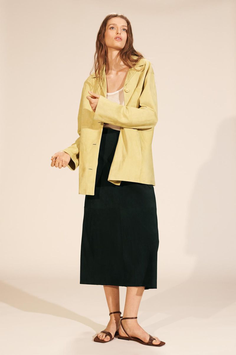 A woman in a yellow blazer and dark skirt poses in a studio. The image has soft lighting and a minimalist aesthetic. Fashion editorial shot.