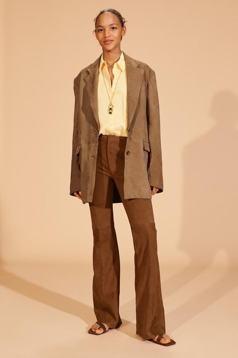 Fashion portrait: A woman in a brown jacket, yellow shirt, and brown pants stands against a neutral backdrop with a soft, diffused light.