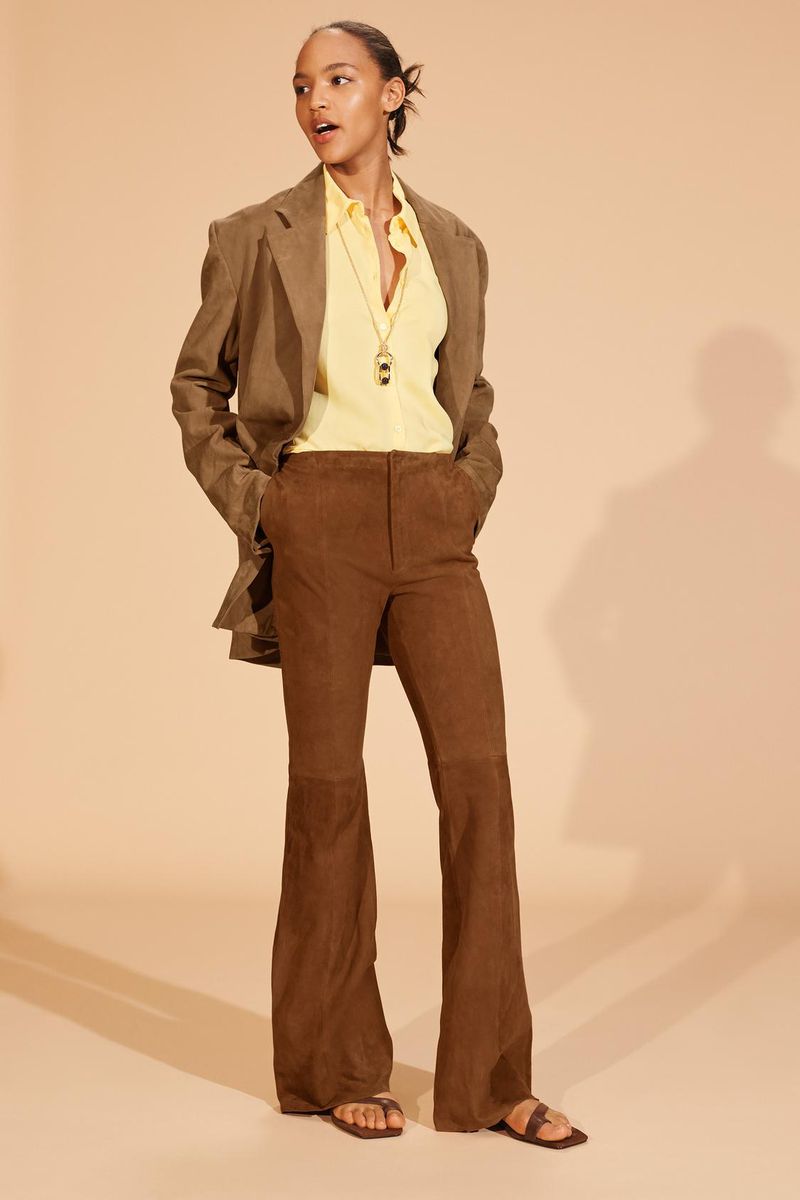 Full shot of a stylish woman in a brown blazer and flared corduroy pants, standing against a light beige backdrop. Fashion editorial.