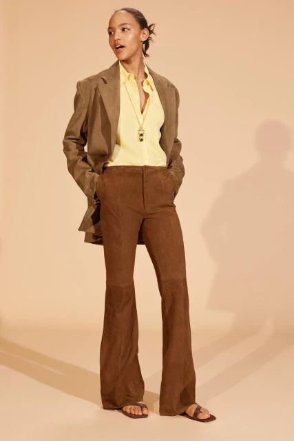 Full shot of a stylish woman in a brown blazer and flared corduroy pants, standing against a light beige backdrop. Fashion editorial.