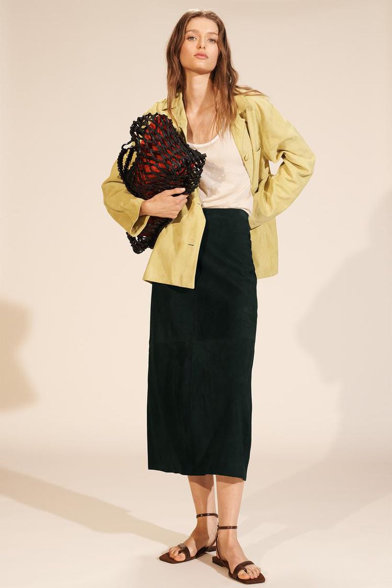 A full shot of a woman wearing a yellow jacket, white top, and green skirt, standing in a studio with a beige backdrop, holding a black string bag.