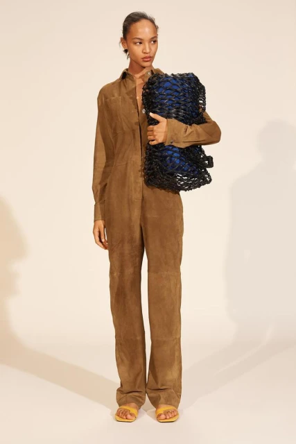 Fashion shot of a tanned-skinned woman posing in a brown jumpsuit with a blue and black woven bag, against a neutral backdrop.