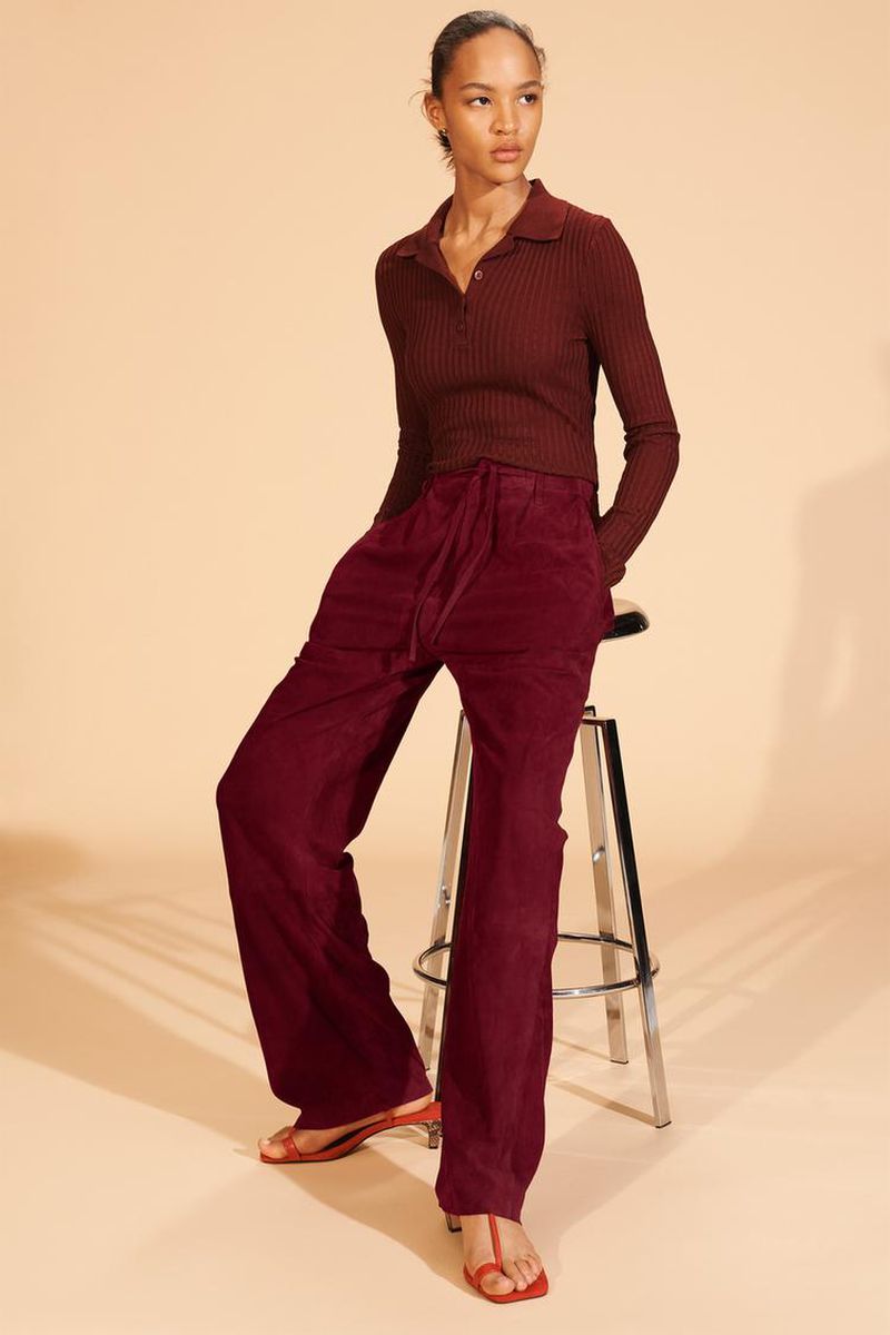 Model in a burgundy long-sleeve polo shirt and matching wide-leg trousers seated on a chrome stool against a warm beige background.