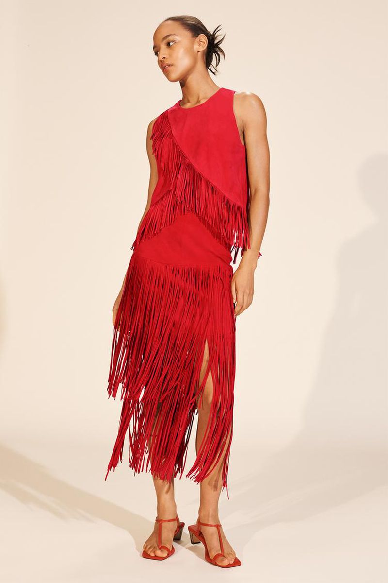 A woman models a red sleeveless top and skirt with fringe details, paired with red heels, against a neutral backdrop in a studio setting.
