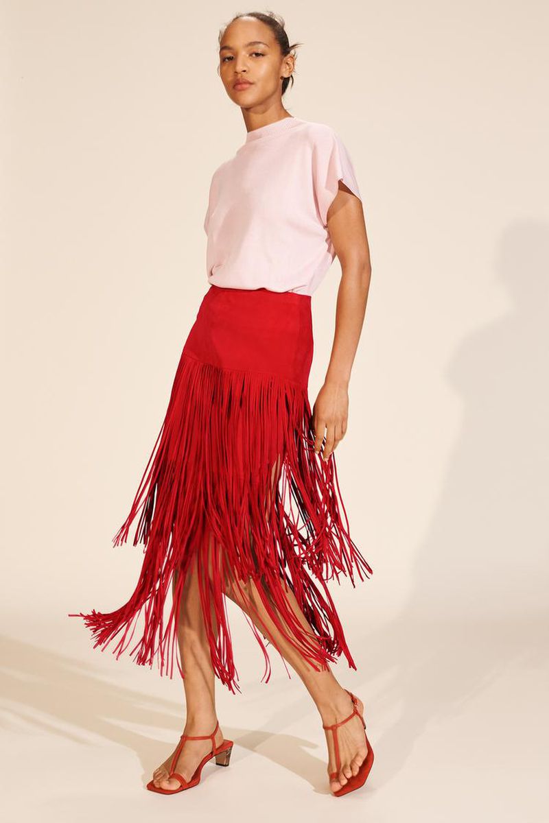 Fashion model wears a pink t-shirt and red fringe skirt with heeled sandals in a studio setting. Fashion editorial content.