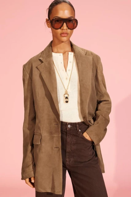 Woman in a taupe blazer, white shirt, brown pants, and sunglasses poses against a pink backdrop in a fashion shot.
