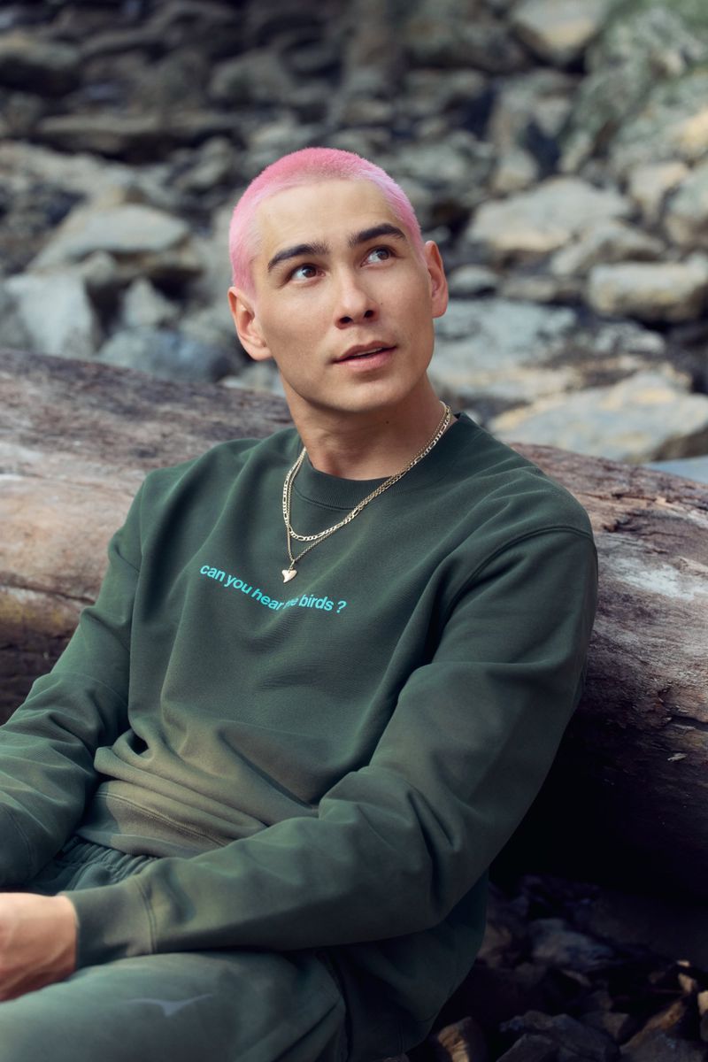 Young man with pink hair in green sweatshirt and sweatpants posing outside against rocky background and fallen tree trunk.
