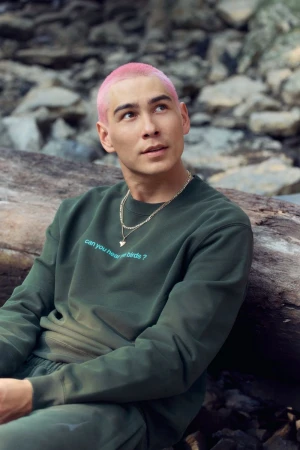 Young man with pink hair in green sweatshirt and sweatpants posing outside against rocky background and fallen tree trunk.
