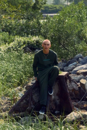 Young adult sits on concrete structure outdoors wearing a matching green sweatshirt and sweatpants in a nature-filled urban setting.