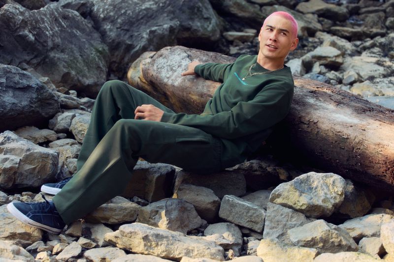 A young adult with pink hair sits on a fallen log surrounded by rocks, wearing a green sweatshirt and pants in an outdoor setting.