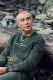 Evan Mock with a pink buzz cut wearing an olive green sweatshirt and gold necklaces, resting against a fallen log outdoors.