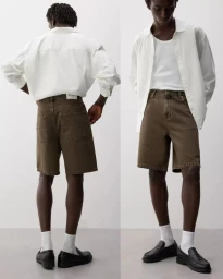 Model wearing a white button-up shirt, brown denim shorts, white socks, and black loafers against a studio backdrop.