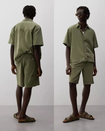 Model Godwin Dekou wearing an olive green short-sleeve shirt and matching shorts with slide sandals against a grey studio backdrop.