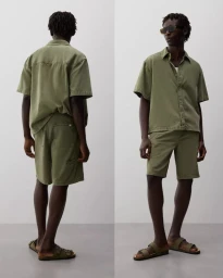 Model Godwin Dekou wearing an olive green short-sleeve shirt and matching shorts with slide sandals against a grey studio backdrop.