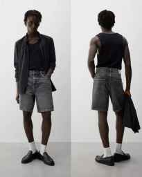 Model wearing a black shirt over a black tank top, grey denim shorts, white socks, and black loafers against a white backdrop