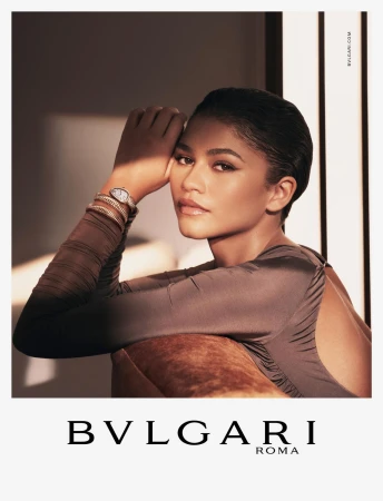 Close-up studio shot of Zendaya wearing Bulgari watch, bracelets, and a sleek taupe dress, set against a soft, neutral backdrop.