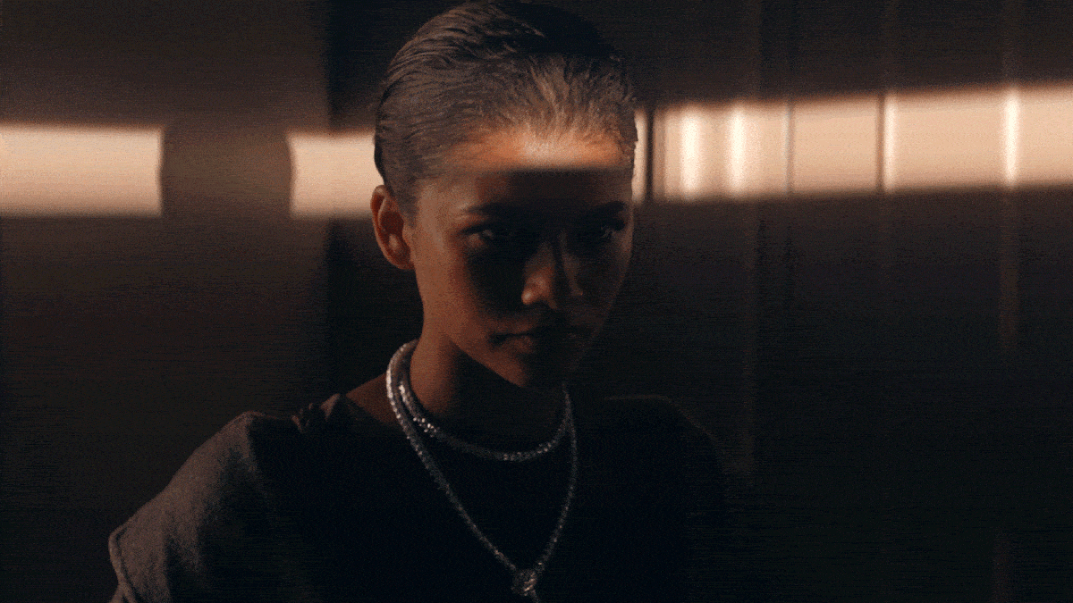 Close-up portrait of a young adult female with sleek dark hair, adorned with a silver necklace, set against a dimly lit indoor background.