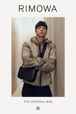Rimowa ad featuring a man wearing a beige jacket and black beanie, posing indoors with a black Rimowa bag on a beige background.