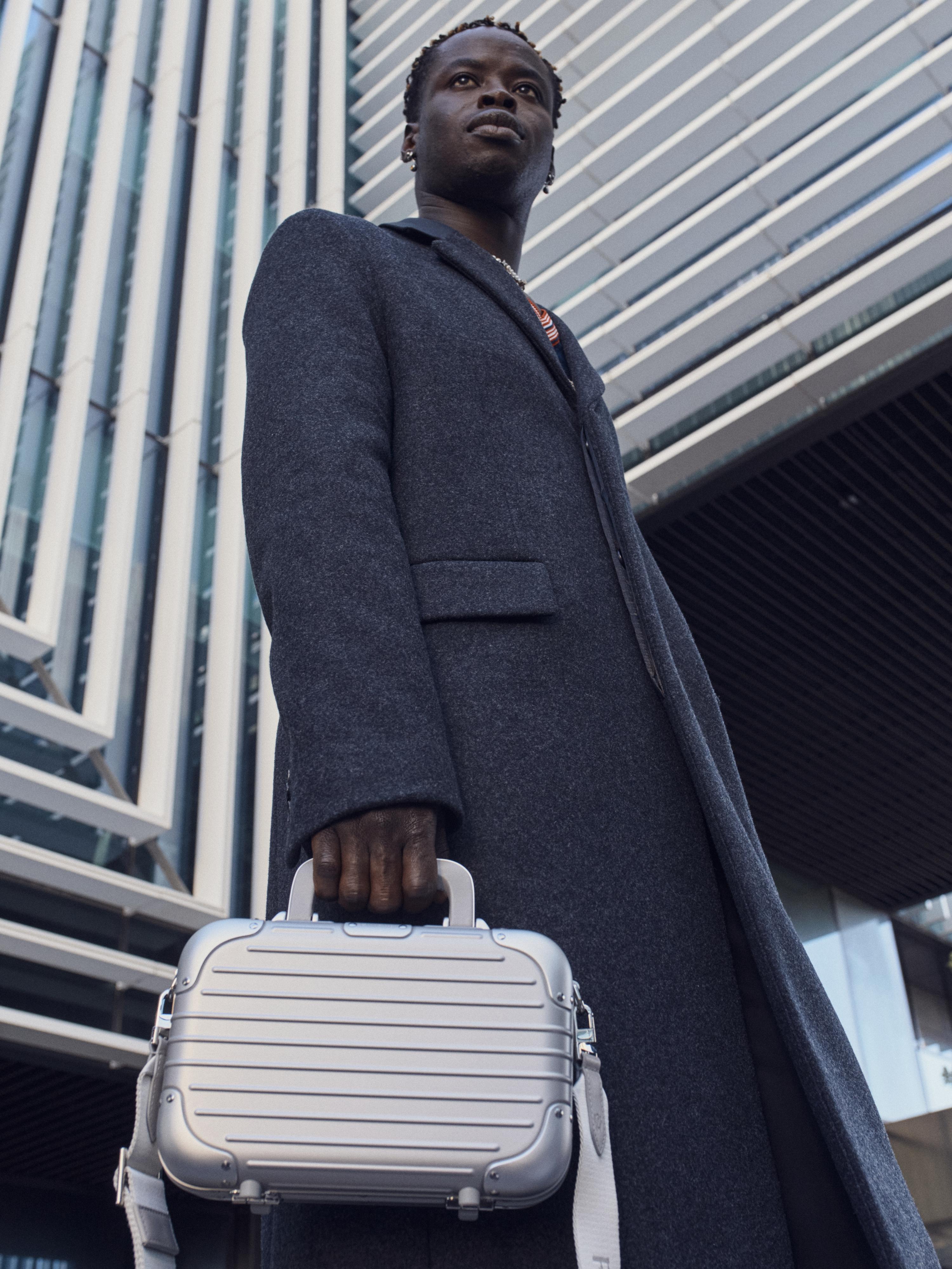 Man in a coat stands outside a modern building holding a silver boxy bag with a shoulder strap, in a low-angle full shot.