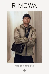 Central Cee wearing a taupe hooded jacket, black beanie, and black RIMOWA Original Bag crossbody against a neutral wall.