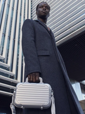 Man wearing a charcoal wool coat holding a silver aluminum mini bag against a modern architectural building backdrop.